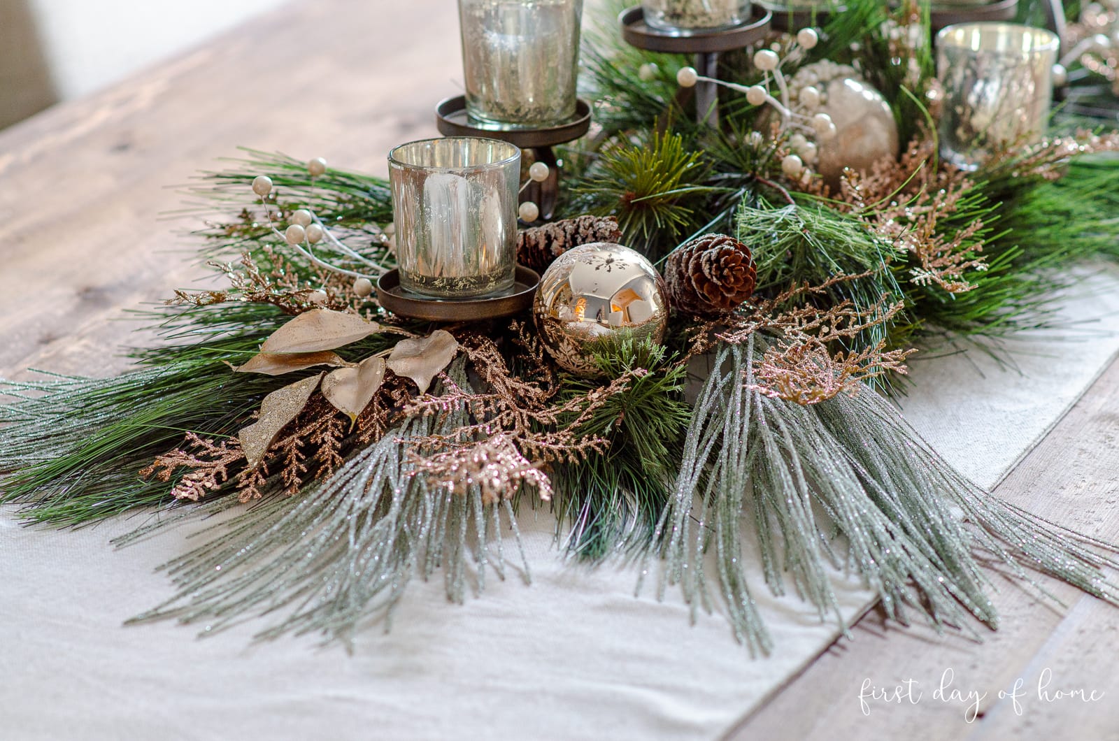 DIY holiday table centerpiece with mixed greenery, mercury glass candleholders, metallic ornaments, and faux floral accents.