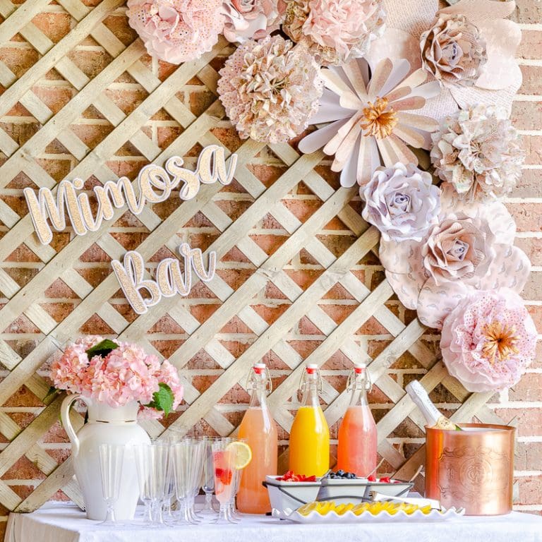 DIY mimosa bar with pink paper flowers and pom poms, fruit juices, champagne flutes and fresh flowers