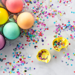 Colored eggs in carton with broken cascron (confetti filled egg) with confetti in background