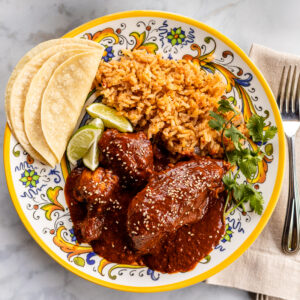 Chicken mole meal with Mexican rice, corn tortillas, and lime wedges served with a bowl of charro beans and salsa