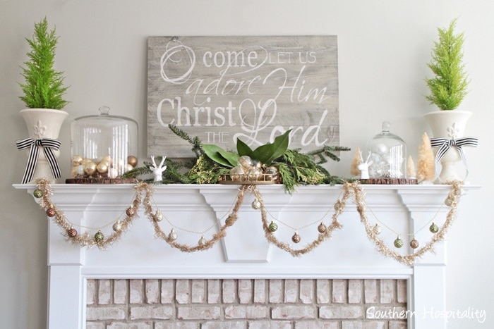 Neutral greige and green Christmas mantel with metallic ornament garland, rustic farmhouse sign, and green topiary trees in urns