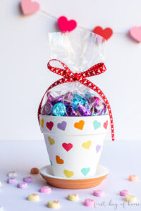 Valentines Day flower pot covered in hearts of various pastel colors. The pot is filled with chocolate candies as a Valentine's Day gift and surrounded by conversation hearts candies.