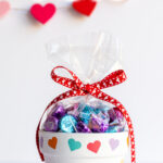 Valentine's Day flower pot with colored hearts painted on it, filled with chocolate kisses and surrounded by conversation heart candy.