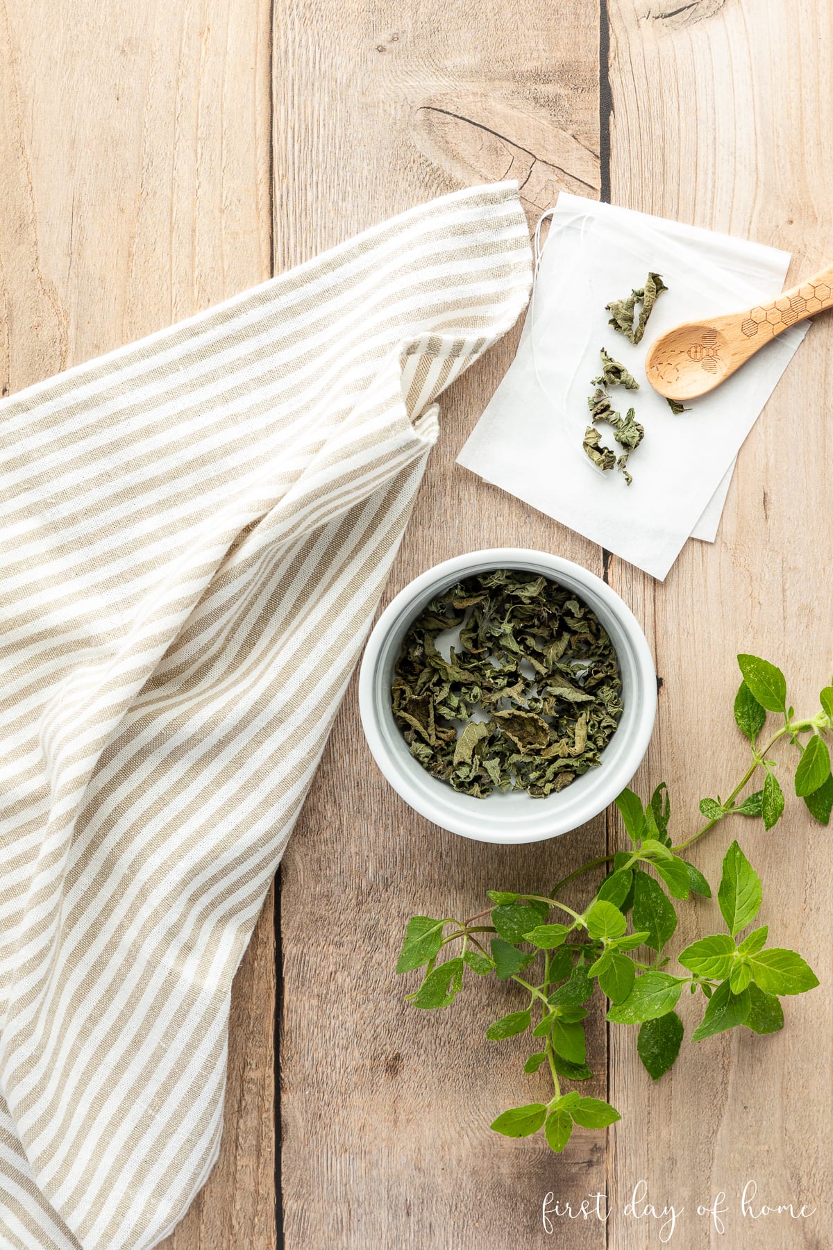 Dried herbal tea leaves shown with fresh herbs, tea bags, wooden spoon, and tea towel