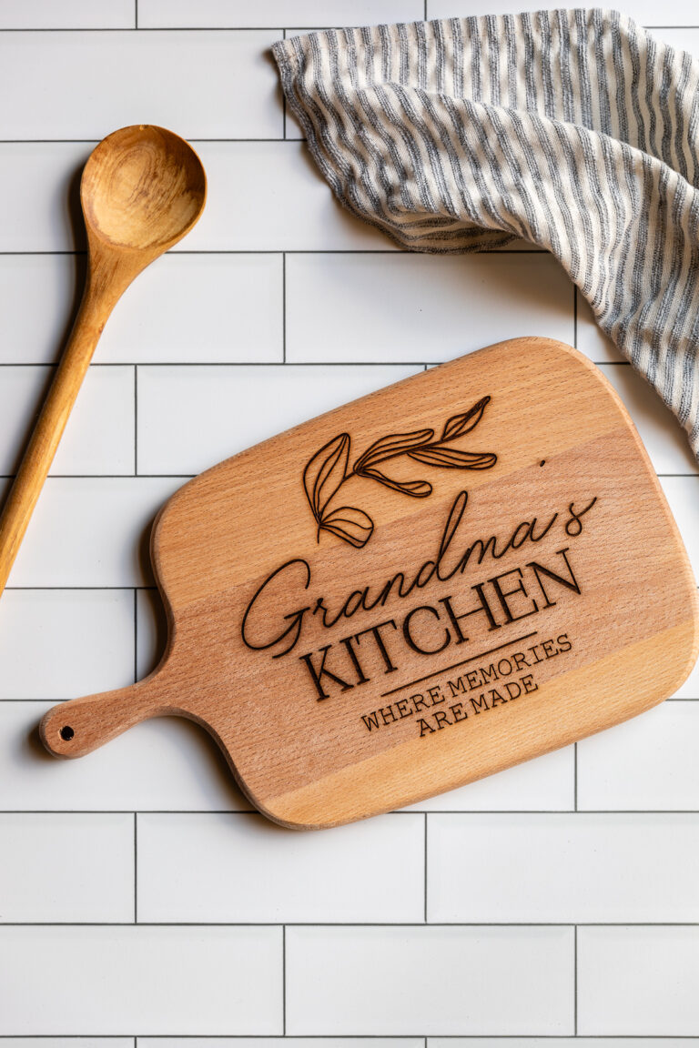 Engraved cutting board that reads "Grandma's Kitchen: Where Memories are Made" shown with wooden spoon and tea towel.