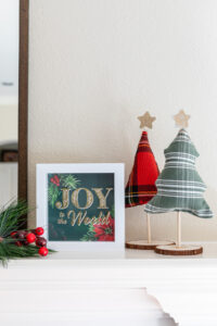 DIY Christmas Shadow Box that reads "Joy to the World" sitting next to two fabric Christmas trees on a fireplace mantel.