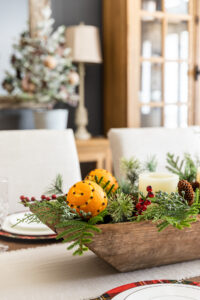 Dough bowl Christmas centerpiece filled with evergreen stems, berries, and orange pomander balls sitting on dining room table.