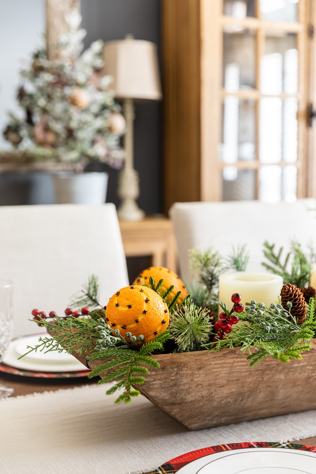 Dough bowl Christmas centerpiece filled with evergreen stems, berries, and orange pomander balls sitting on dining room table.
