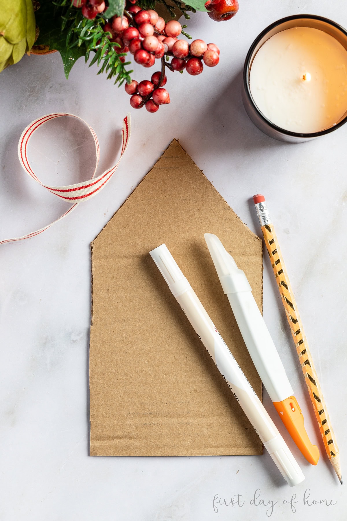 Cardboard house cutout with white paint pen, craft knife, and pencil, shown with a candle and ribbon.