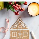 Cardboard gingerbread house shown with candle, greenery, and greenery.
