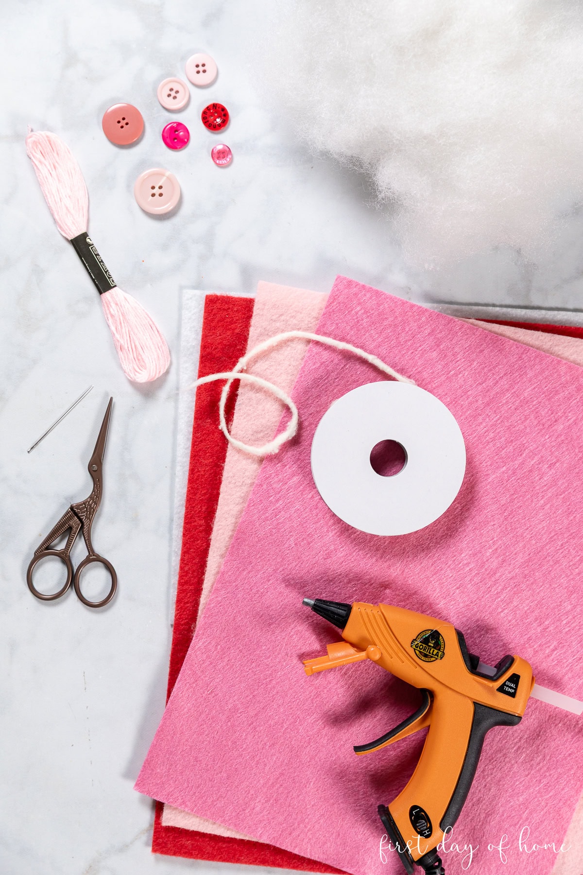 Felt heart ornament supplies, including felt, embroidery floss, scissors, ribbon, buttons, and hot glue.