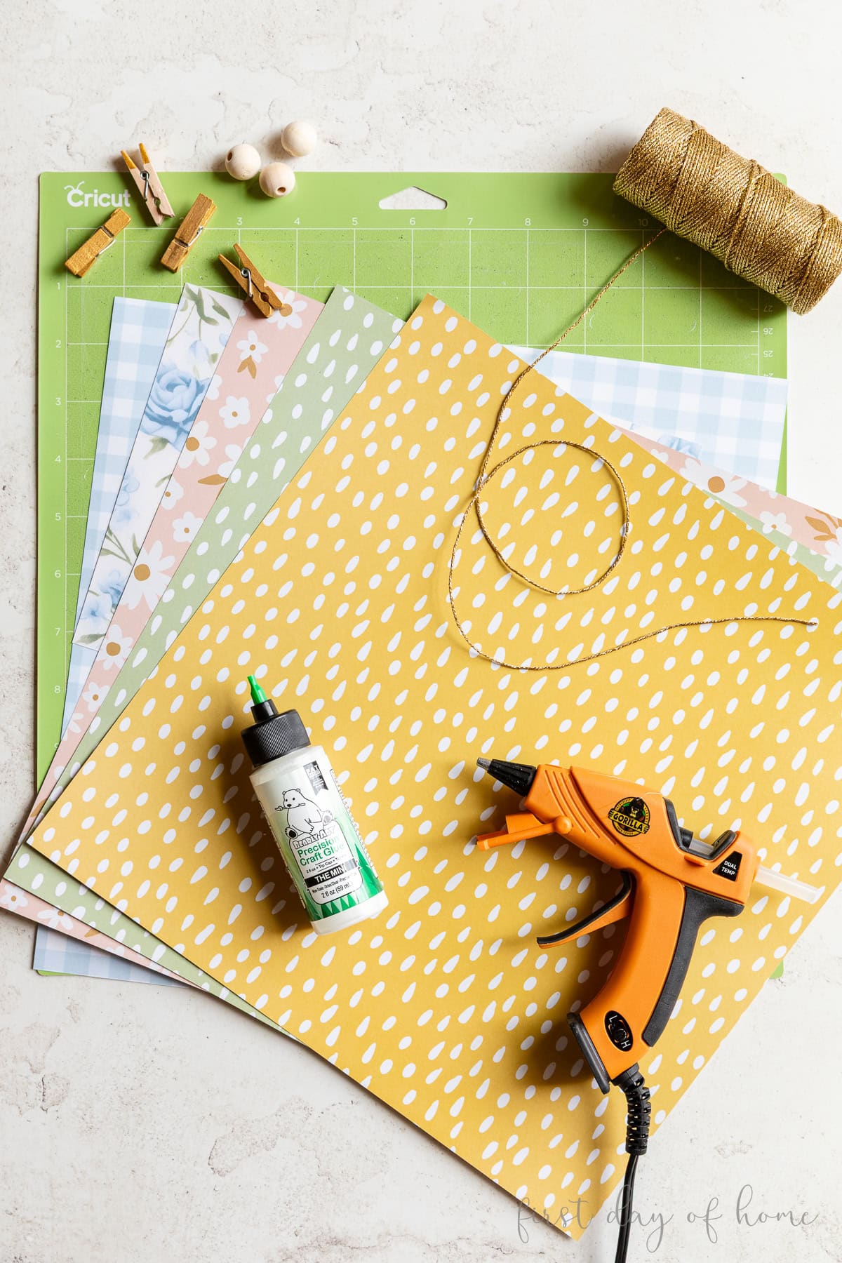 Supplies for making DIY paper Easter ornaments, including scarpbook paper, craft glue, hot glue, ribbon, clothespins, wood beads, and a Cricut cutting mat.