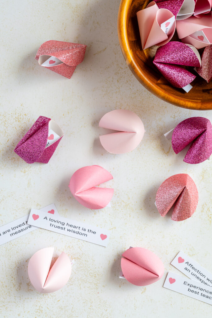 Paper fortune cookies in a bowl and scattered with paper fortunes.
