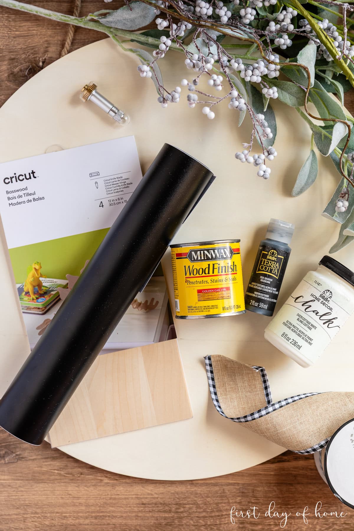 Supplies to make a DIY wooden door hanger, including an unfinished wood sign, iron-on vinyl, basswood, wood stain, paint, and faux floral stems.